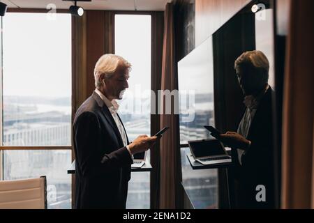 Side view of senior businessman using smart phone while standing by reflection on television in board room Stock Photo