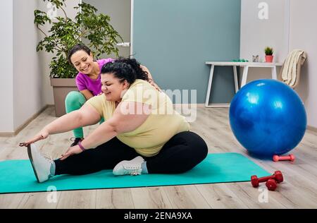 Overweight woman exercises with a personal trainer. Brunette plus-size doing stretching workout on a fitness mat Stock Photo