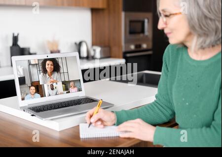 Back view of smiling entrepreneur, aged woman, multiethnic business team, writing notes, on the kitchen, brainstorming, looking for new directions for promotion, on distance, writes down ideas Stock Photo