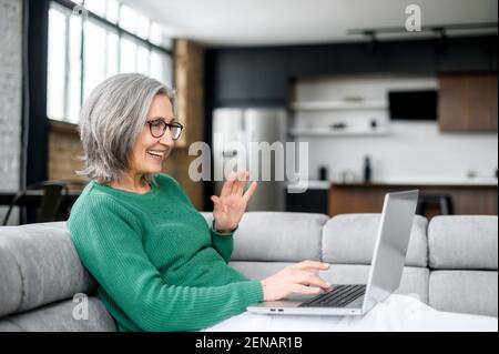 Video call concept, lovely elderly woman with nice wrinkles, narrow lips, smiling, chatting with family, using a laptop for video connection, remote meeting, say hi, glad to see, lying on the couch Stock Photo