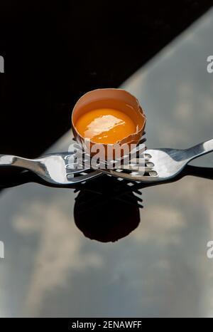 Broken fresh chicken egg (Hen egg) balanced on a composition of two intertwined forks over black floor. Copy space, Selective focus. Stock Photo