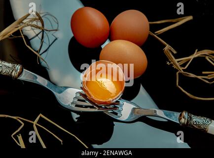 Broken fresh chicken egg (Hen egg) balanced on a composition of two intertwined forks and Three fresh chicken eggs over black floor. Selective focus. Stock Photo