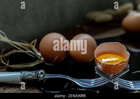 Broken fresh chicken egg (Hen egg) balanced on a composition of two intertwined forks and Two fresh chicken eggs over black floor. Selective focus. Stock Photo