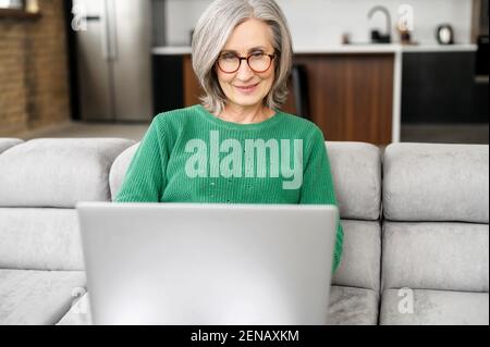 Happy elderly woman sit relax on couch in living room, work on laptop, smiling, modern mature 60s female enjoy leisure weekend at home, browsing wireless internet on computer gadget Stock Photo