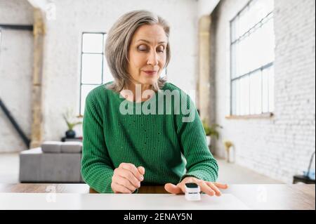 Aged healthy woman with wrinkles on the face, dressed in green warm sweater, determines the saturation of the lungs with oxygen, at home, loft style apartment, on the isolation,caring about her health Stock Photo