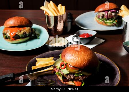 Different burgers on the table, served frenc fries and sauces on plates. The closest spicy beef burger with jalapenos, pickled cucumbers and avocado. Stock Photo
