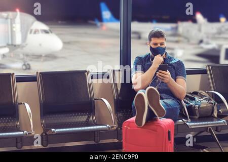 Man wearing face mask sitting in airport departure terminal using mobile phone tracking app while waiting for boarding Stock Photo