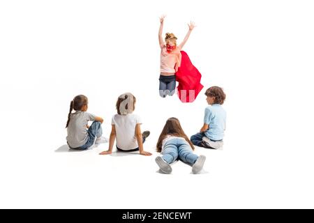 Power. Child pretending to be a superhero with his friends sitting around  him. Kids excited, inspired by their strong friend in red coat isolated on  white background. Dreams, emotions concept Stock Photo 