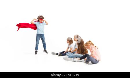 Rescuer. Child pretending to be a superhero with his friends sitting around him. Kids excited, inspired by their brave friend in red coat isolated on white background. Dreams, emotions concept. Stock Photo
