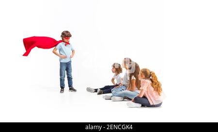 Rescuer. Child pretending to be a superhero with his friends sitting around him. Kids excited, inspired by their brave friend in red coat isolated on white background. Dreams, emotions concept. Stock Photo