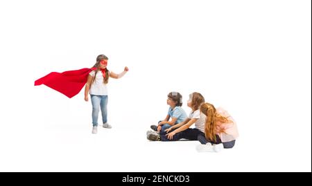 Flying. Child pretending to be a superhero with his friends sitting around  him. Kids excited and inspired by their brave friend in red coat isolated  on white background. Dreams, emotions concept Stock