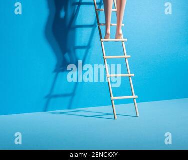 Doll legs on a stepladder on blue background. Success and progress concept. Minimal composition. Stock Photo