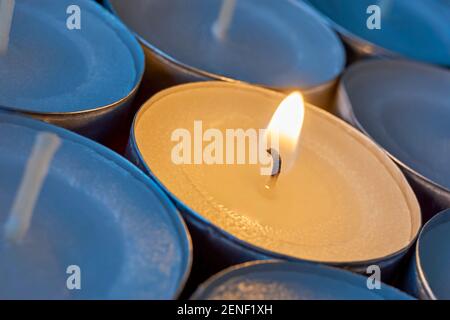 Closeup of tea lights with one being lit. A single warm orange flame between  candles in blue shadow. Christmas, condolences or funeral background. Stock Photo