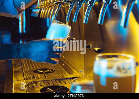 Young man bartender pouring lager beer from tap to glass in neon light. Stock Photo