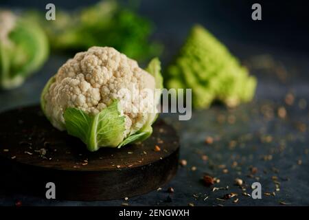 closeup of a raw baby cauliflower head in a wooden plate, on a dark stone surface sprinkled with different seeds and spices and a baby romanesco brocc Stock Photo