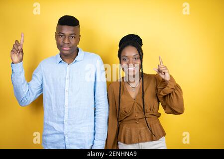 Young african american couple standing over yellow background amazed and surprised looking at the camera and pointing up with fingers and raised arms Stock Photo