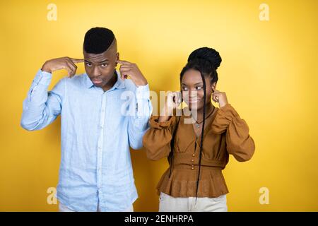 Young african american couple standing over yellow background covering ears with fingers with annoyed expression for the noise of loud music. Deaf con Stock Photo