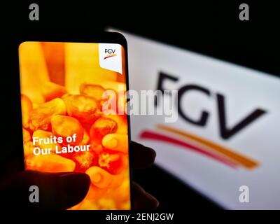 Person holding cellphone with logo of Malaysian agriculture company FGV Holdings on screen in front of webpage. Focus on center of phone display. Stock Photo