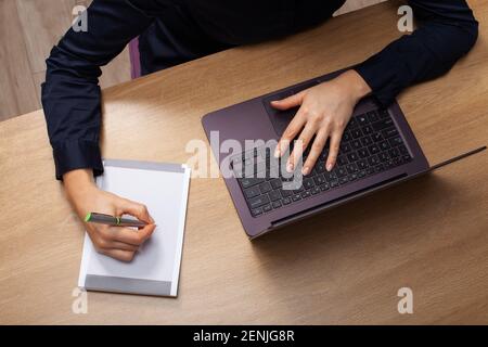 Detail top view of a woman at home sitting at the table with a laptop, taking notes in a notebook. Dressed in a black shirt. Stock Photo