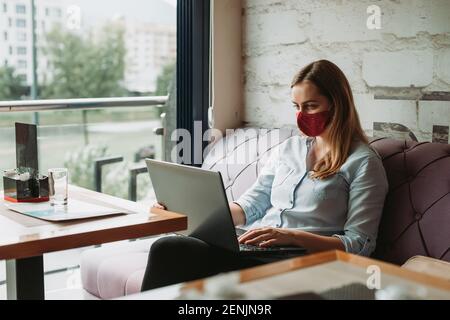Woman freelancer indoors with face mask working on laptop in coffee shop, remote work Stock Photo