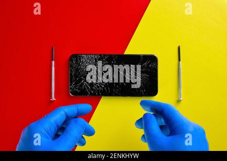 The phone with the broken screen lies on a red and yellow background with small screwdrivers and the hands of a master Stock Photo