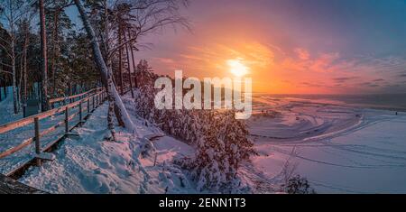 Colorful sunset over snowy sea coast and pine forest with wooden boardwalk path. Beautiful view of Baltic sea coast with snow-covered fir, spruce Stock Photo