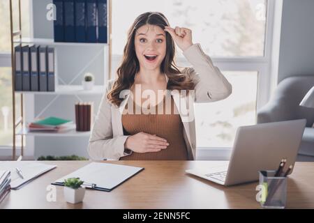 Photo portrait of pregnant excited woman lifting up glasses sitting at desk in modern office Stock Photo