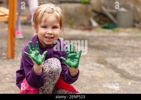 Young happy child, joyful cheerful little school age girl showing both of her hands covered in green paint, outdoors portrait, copy space, lifestyle s Stock Photo