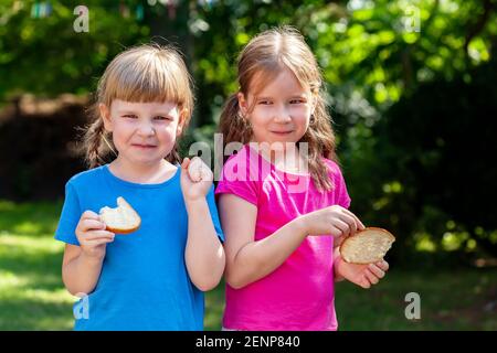 Two happy little girls eating slices of bread with butter smiling, outdoors portrait. Kids, sisters, children eating food outside, lifestyle shot, sun Stock Photo