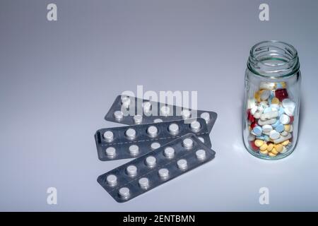 Blister pack of white pills next to a transparent glass bottle containing yellow, blue, red, and white tablets and capsules. Stock Photo