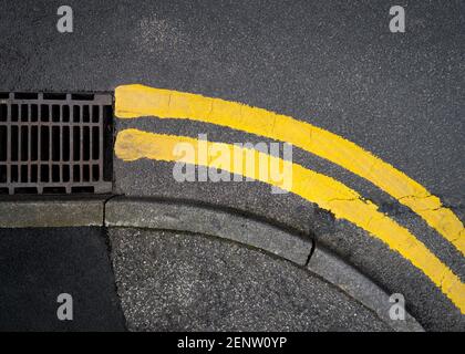 No parking two double yellow lines restriction painted on black tarmac road at junction corner to prevent cars trying to park up to metal drainage man Stock Photo