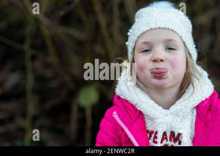 Little girl in pink winter coat and white woolie hat sticking out her tongue and blowing raspberries, little one being cheeky, kid being cheeky Stock Photo