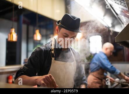 Young guy in hat and apron cooking steamy dish during work in restaurant Stock Photo