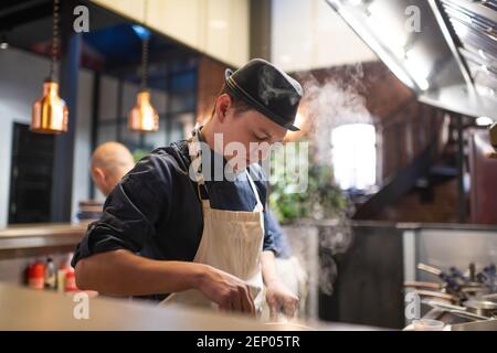 Young guy in hat and apron mixing dish in saucepan while working in cafe kitchen Stock Photo