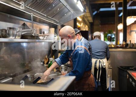 Bald man using tongs to mix pasta on frying pan while cooking in kitchen of cafe Stock Photo