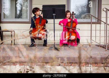 Two children sit outside in fancy dress playing cellos on porch Stock Photo