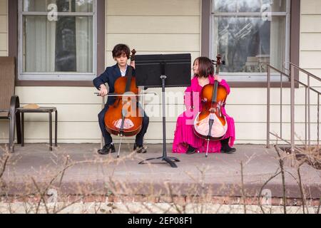 A tween boy sits on porch playing cello in suit while sister watches