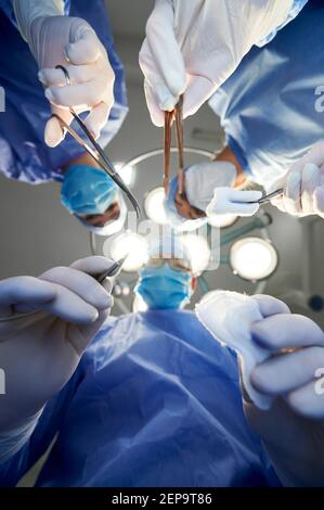 View from below, focus on doctors hands holding scalpel, scissors and forceps with tampon while looking at patient during surgery in operating room at hospital. Concept of medicine and surgery. Stock Photo