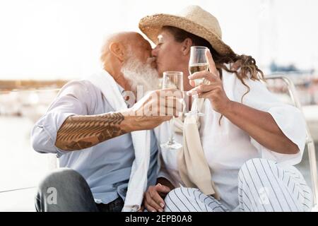Senior couple kissing and cheering with champagne on boat during summer vacation - Focus on glasses