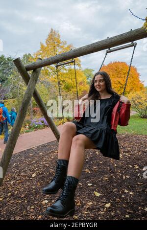 Young dark-haired female in a short black dress and big boots swinging on a swing in an autumn park in Berlin Stock Photo