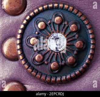 Macro shot of a copper medaillon with flower pattern and a light reflection on the glass bead in the centre. Fake purple leather background Stock Photo
