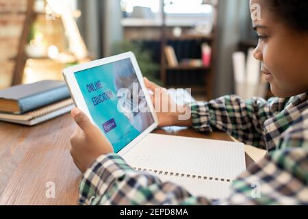 Side view of African elementary schoolboy looking at display of touchpad while sitting by table in living-room and watching online lesson Stock Photo