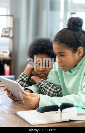 Mixed-race siblings with digital tablet sitting by table in living-room and watching online educational video while preparing homework Stock Photo