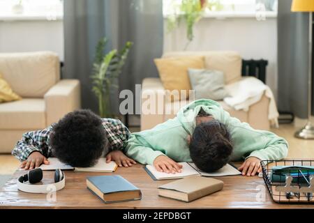 Tired elementary schoolchildren in casualwear lying on their open copybooks on table during preparation of homework in living-room Stock Photo