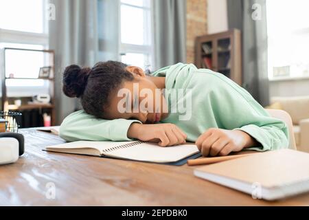 Tired mixed-race schoolgirl in hoodie keeping head on open copybook on table during preparation of homework in living-room against windows Stock Photo