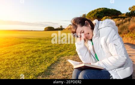 Dreamy photo of middle aged woman reading a book while sitting on a nice grassy area, during the sunset. Stock Photo