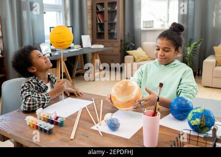 Two adorable mixed-race elementary schoolkids making planets while preparing for lesson of astronomy by table in home environment Stock Photo