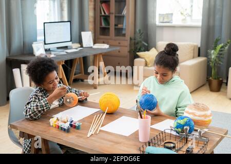 Two adorable siblings of elementary age painting planets while sitting by table in home environment and preparing for the lesson of astronomy Stock Photo