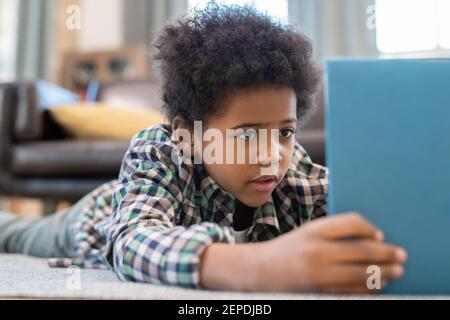 Cute elementary schoolboy with digital tablet lying on the floor against black leather couch in living-room and watching curious online video Stock Photo