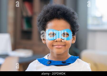 Cute mixed-race boy in costume of superhero looking at you through funny blue paper eyeglasses while standing in front of camera Stock Photo